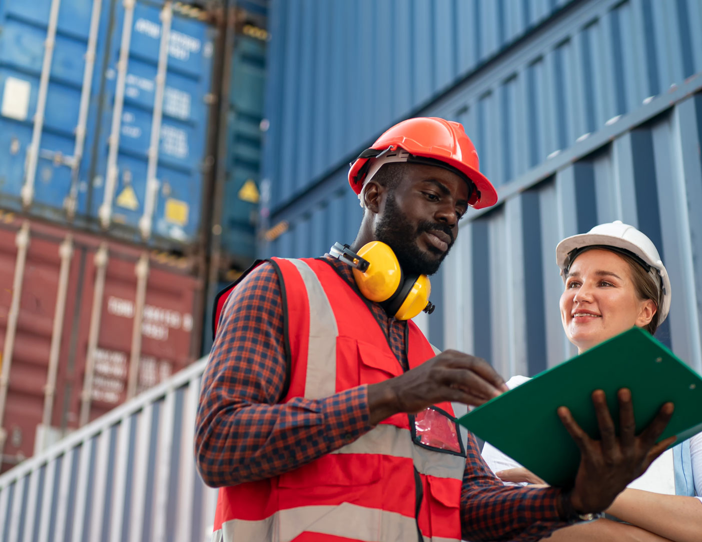 A man and woman wearing hard has look over paperwork while standing next to stacked shipping containers