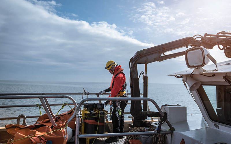 A man in safety equipment examines research equipment on the deck of a vessel.