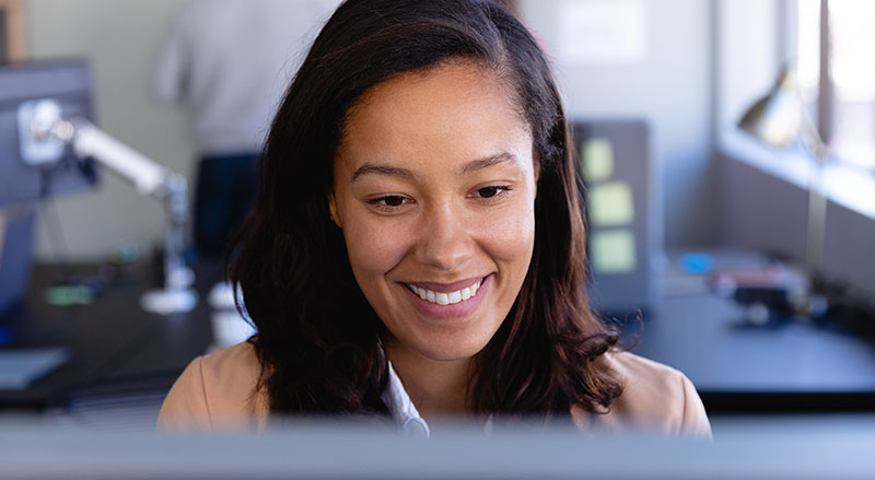 A woman smile at a computer screen. People work on a whiteboard behind her.